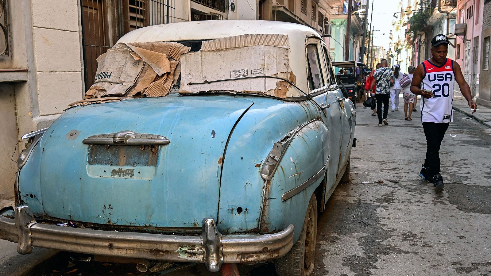 Un hombre con una camiseta de EE.UU. camina cerca de un coche viejo en una calle de La Habana