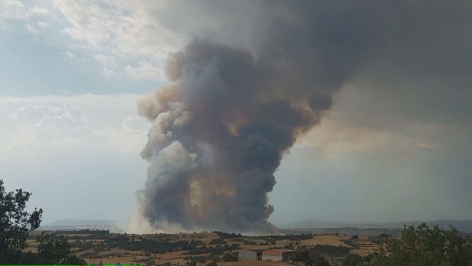 Gran columna de humo gris y marrón, similar a un hongo, sobre campos de cultivo.  Cielo con nubes y humo.  Pirocúmulo.