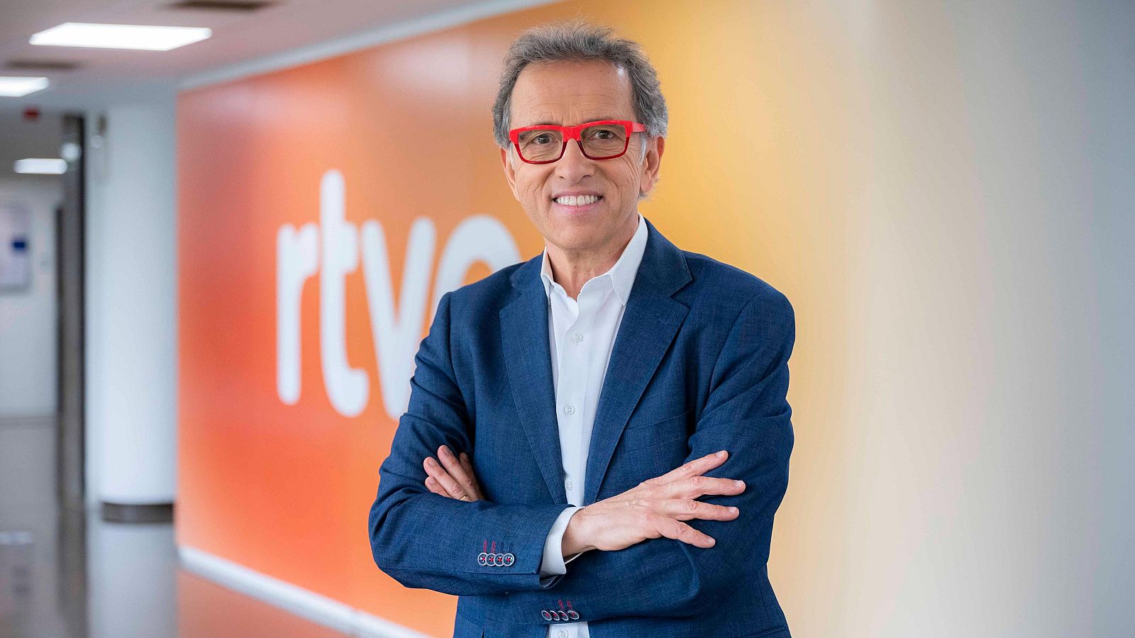 Jordi Hurtado sonríe, con americana azul marino y gafas rojas, frente al logo de RTVE.