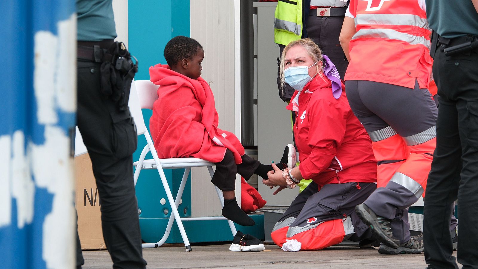 Voluntaria de Cruz Roja asiste a un niño en una silla plegable; otros miembros de la organización y agentes de autoridad presentes.  Atención a menores en un puerto.