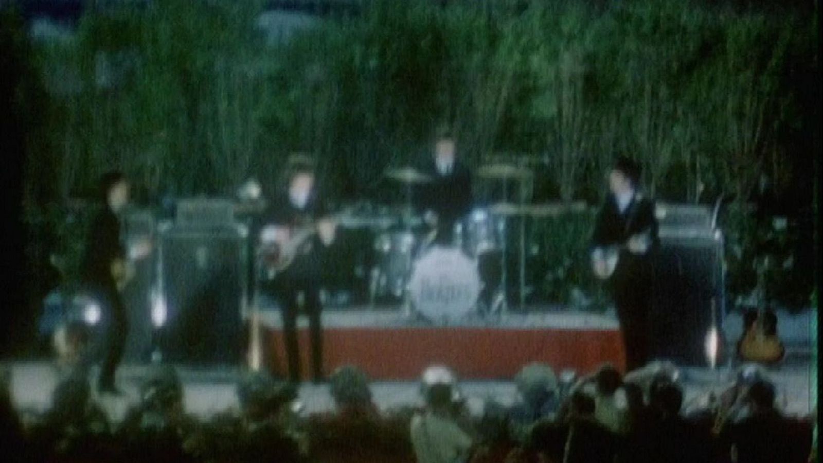 Escena de concierto: cuatro músicos en un escenario rojo tocando instrumentos ante una gran audiencia. Imagen de baja calidad.