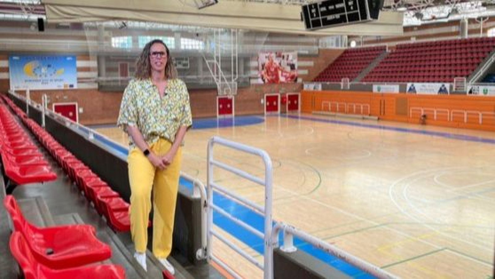 Exjugadora de baloncesto en un pabellón, vestida con camisa floral y pantalón amarillo, posa en las gradas con expresión neutra mirando a cámara. Pista vacía.