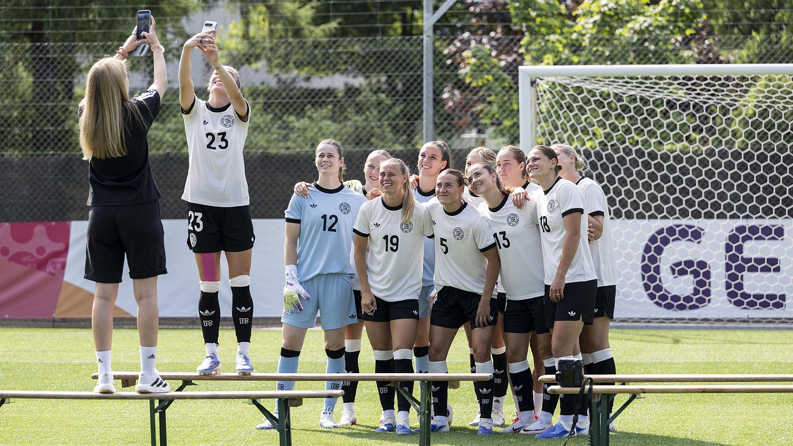 Selfie de jugadoras de fútbol en entrenamiento, con equipaciones blancas y negras, espinilleras y una portería al fondo.