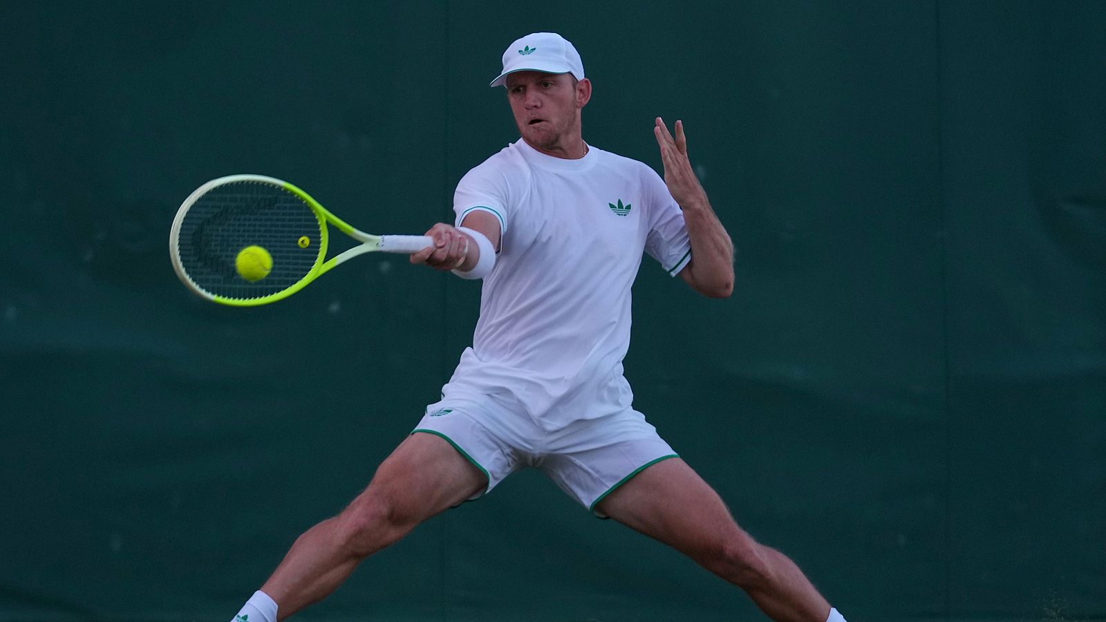 Tenista en plena acción durante un partido, con ropa deportiva blanca y detalles verdes, impactando la pelota con su raqueta. Fondo verde de pista de tenis.