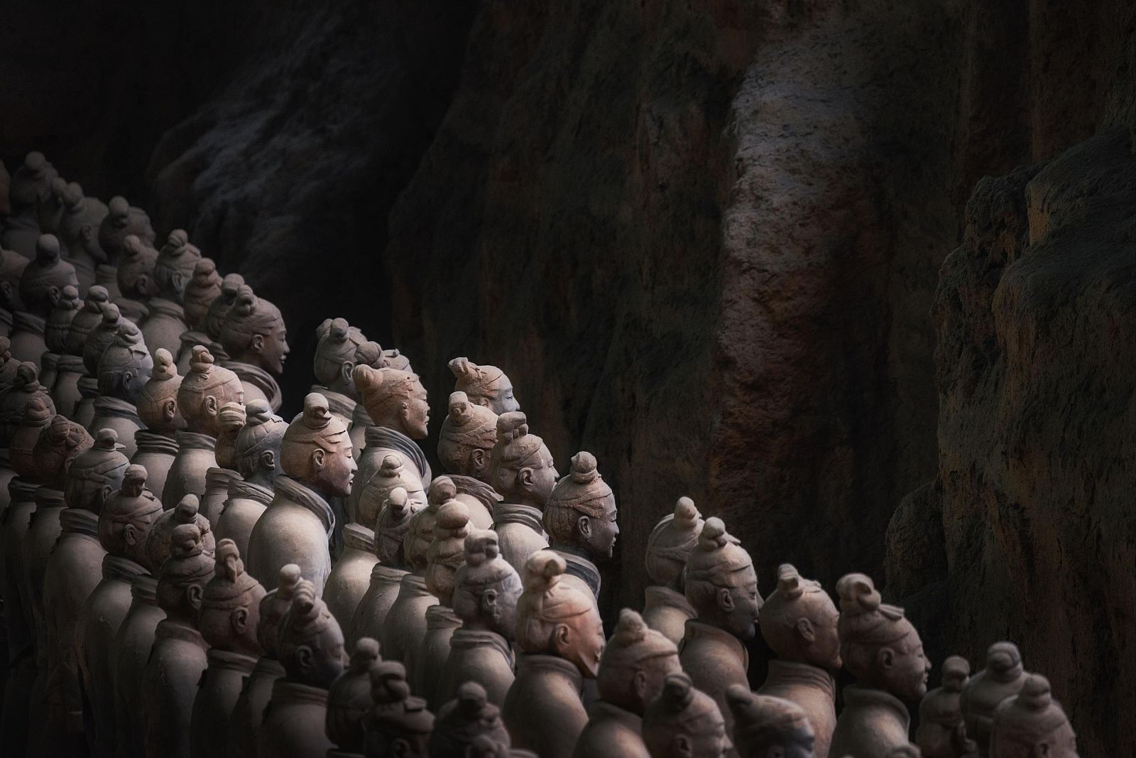 Numerosos guerreros de terracota, de tamaño natural y con rasgos faciales únicos, dispuestos en filas dentro de una fosa. Iluminación tenue que resalta detalles de las esculturas.