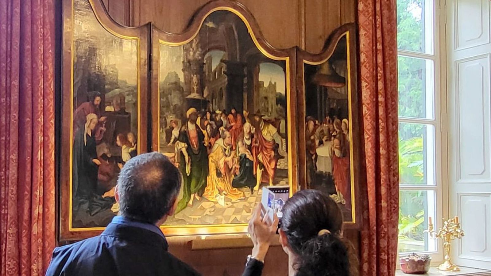 Tríptico religioso recuperado, observado por dos personas, una de ellas fotografiándolo.  Ambiente interior con cortinas rojas y decoración dorada.