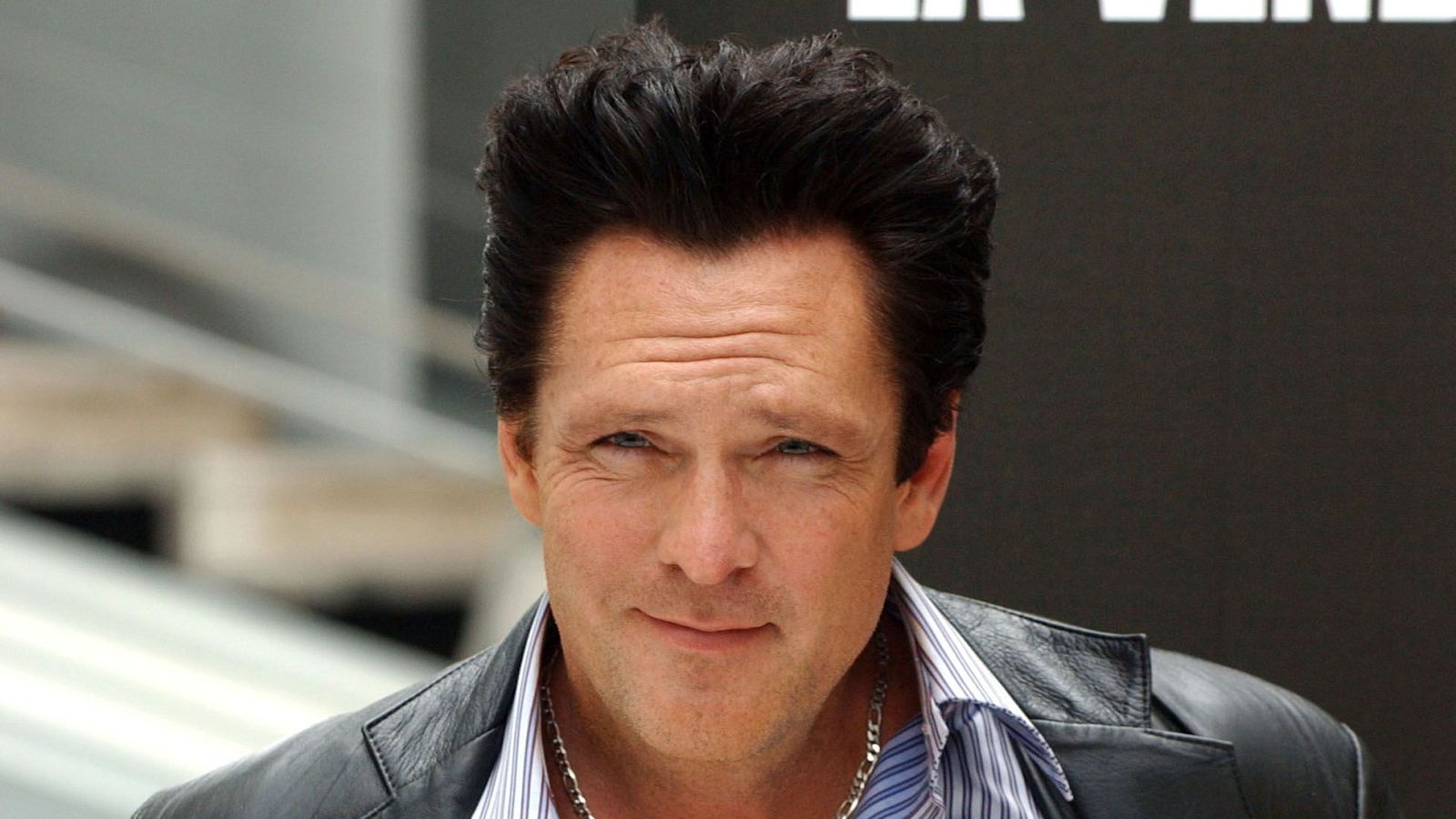 Retrato de Michael Madsen con camisa a rayas, chaqueta de cuero negra y cadena plateada; expresión serena. Falleció a los 67 años.