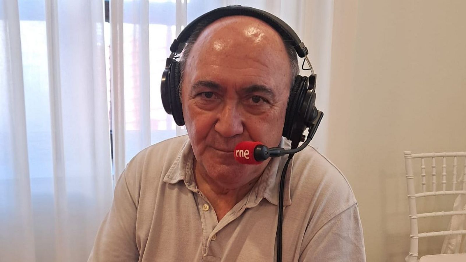 Hombre de mediana edad con auriculares y micrófono 'rne' en una habitación con cortina blanca, posiblemente grabando o en una entrevista.