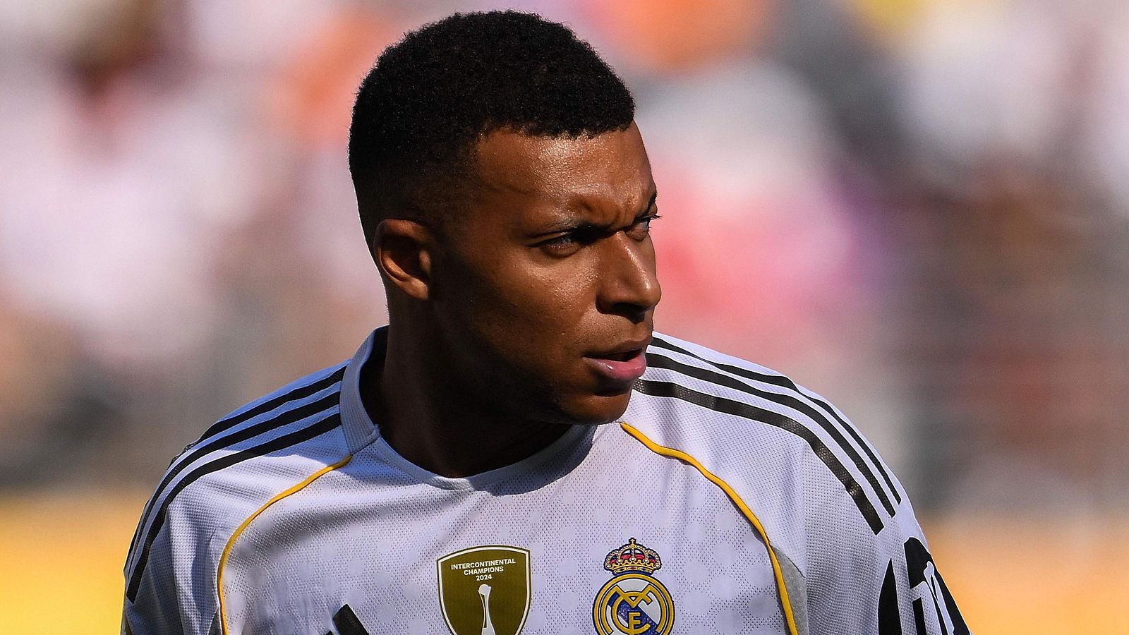 Futbolista con camiseta blanca del Real Madrid, escudo visible y parche dorado.  Expresión seria, fondo desenfocado con tonos naranja, blanco y rosa.