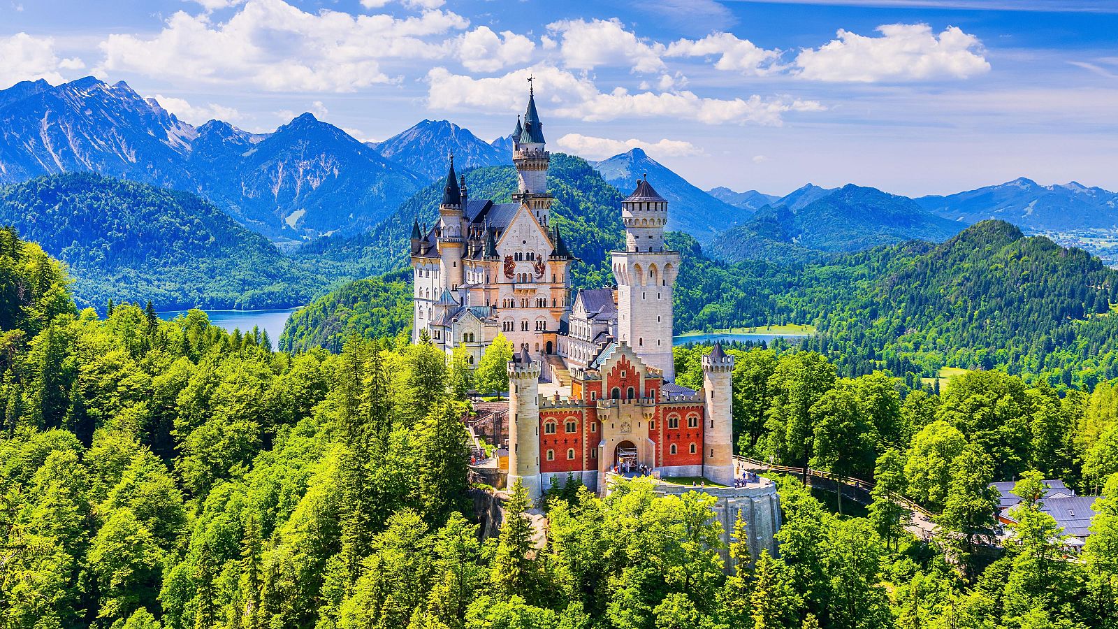 Majestuoso castillo de cuento de hadas en paisaje montañoso, con torres de diversas alturas y colores blanco, beige y rojizo.  Se observa un lago en el fondo.