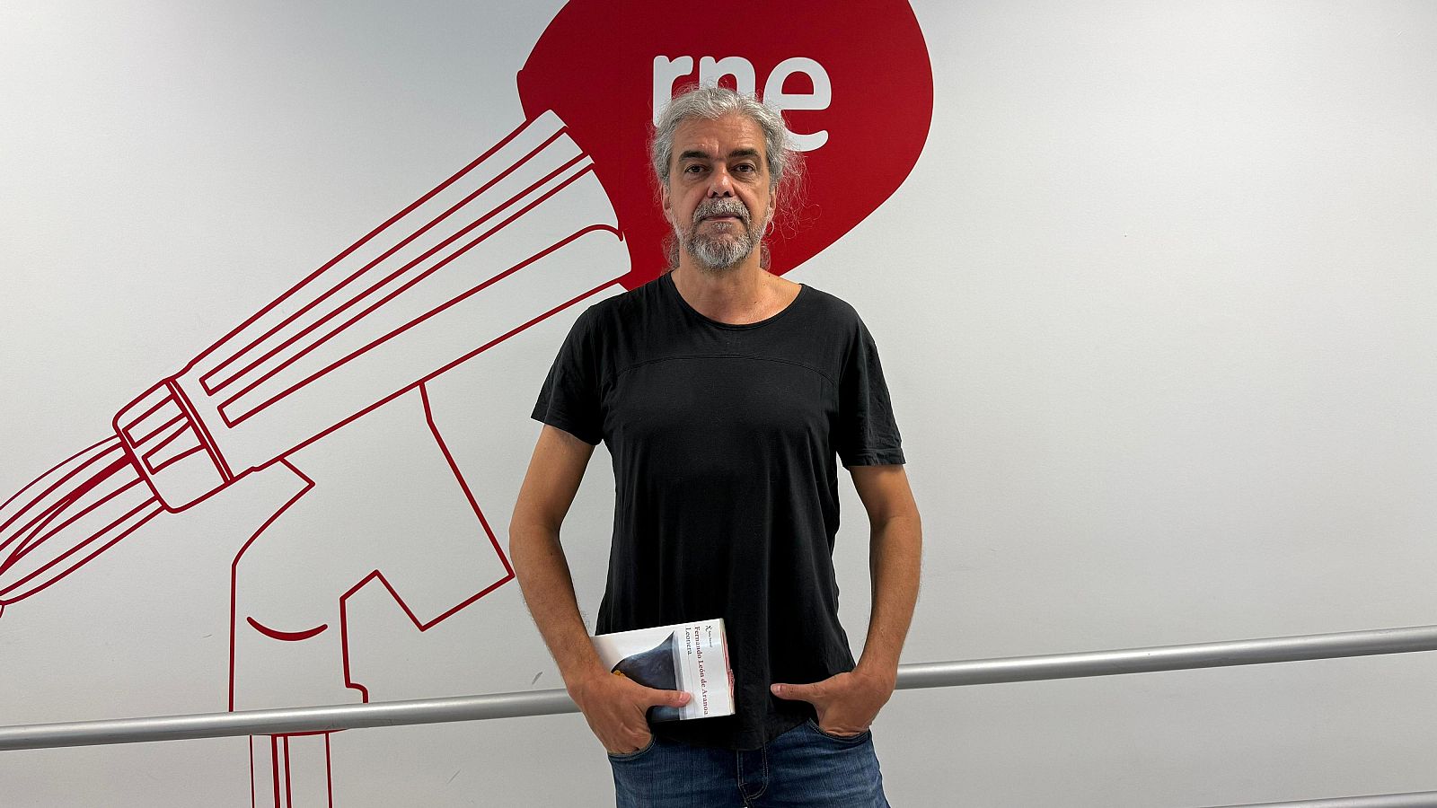 Hombre de pelo largo y canoso con barba, camiseta negra y vaqueros, sostiene un libro en un pasillo de RNE frente a un logo rojo de la emisora.