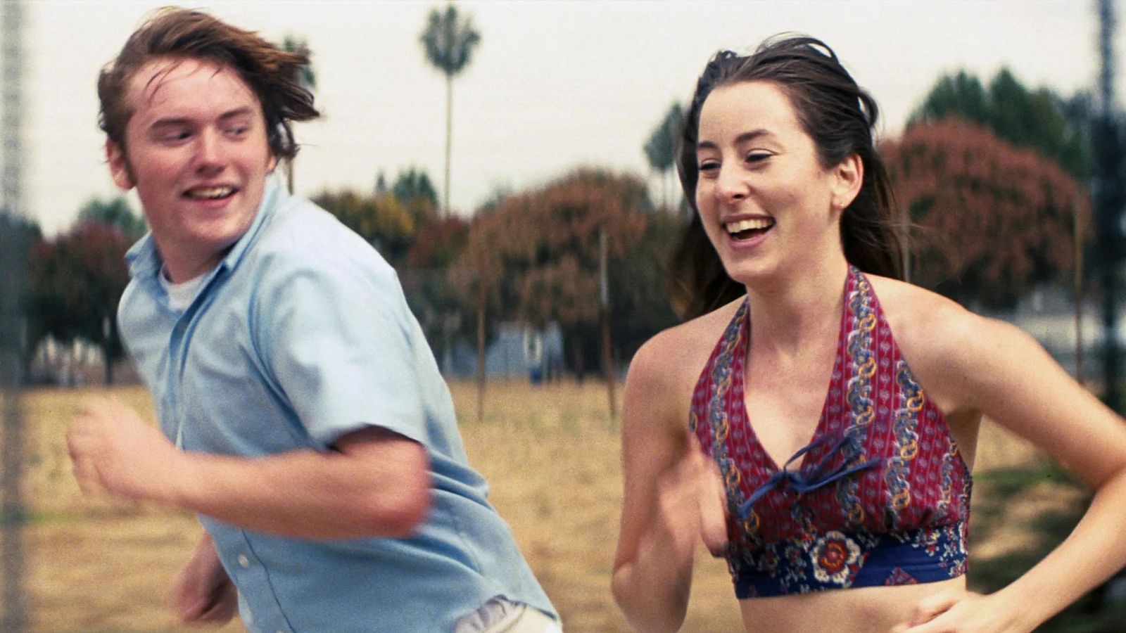 Escena soleada de 'Licorice Pizza' con Cooper Hoffman y Alana Haim corriendo y sonriendo; él con camisa azul, ella con top floral.