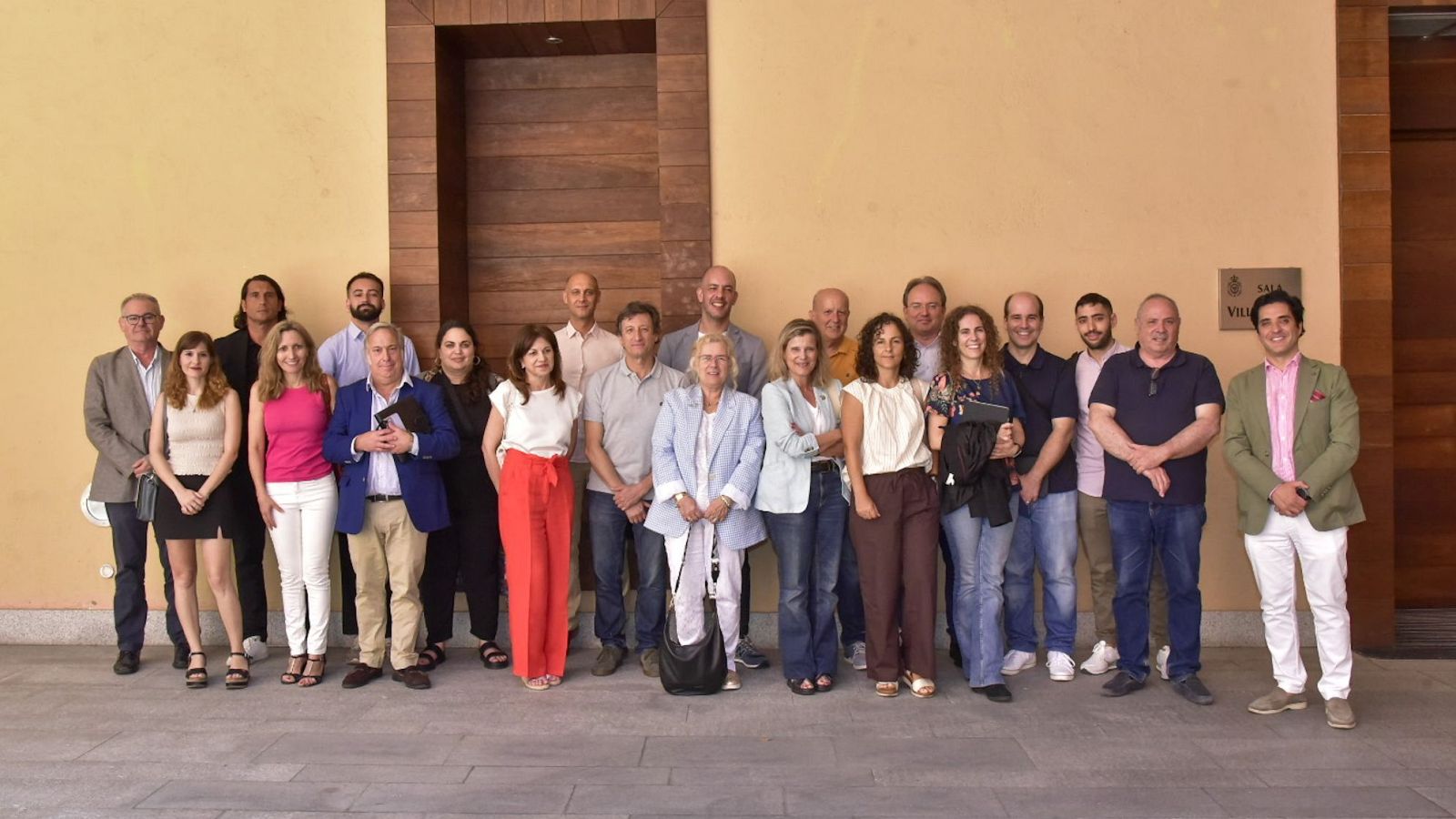 Foto de grupo de unas 20 personas con ropa informal y de negocios, reunidas en una jornada de trabajo frente a una pared beige y una puerta de madera.