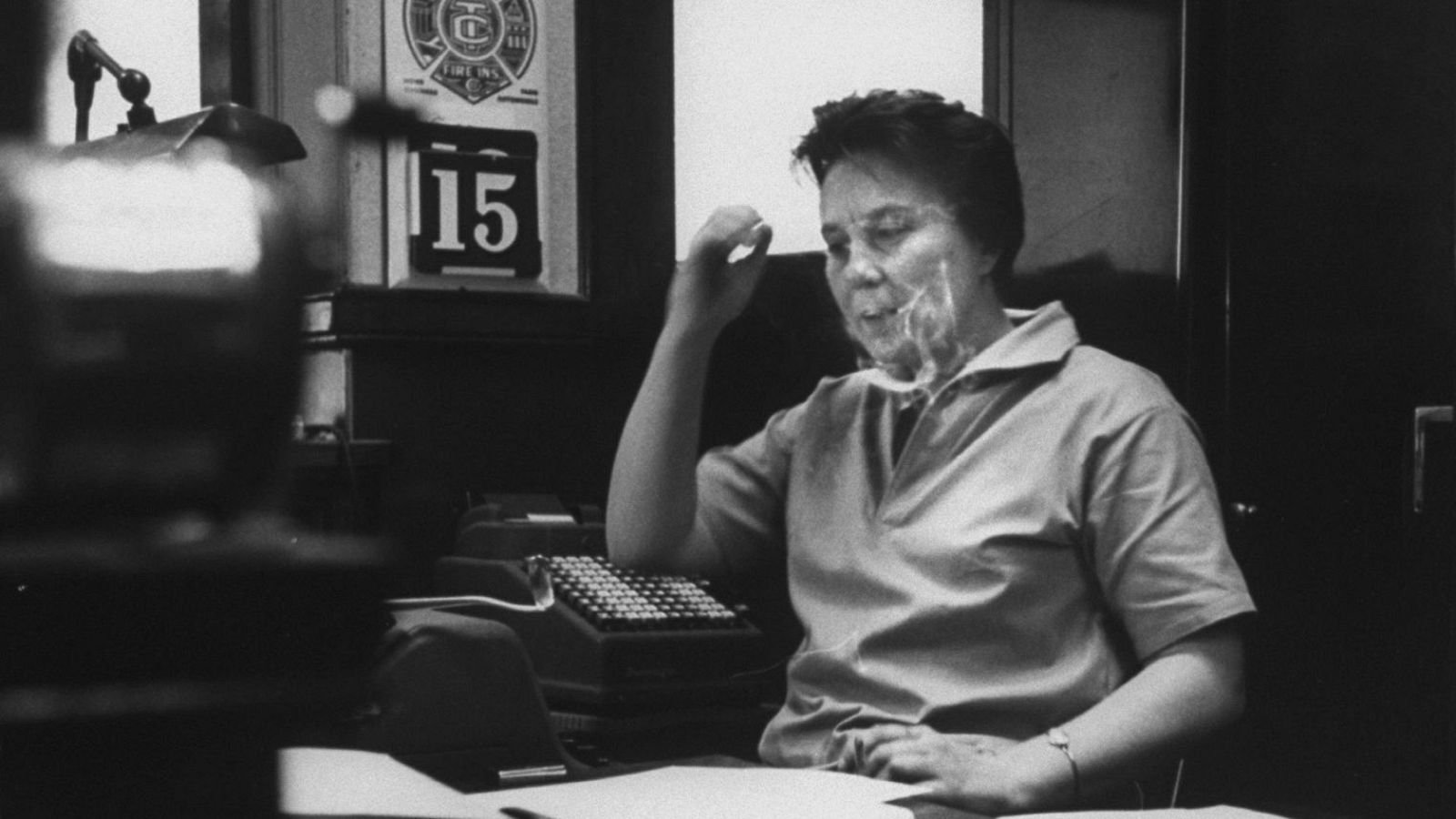 Mujer de mediana edad fumando en una oficina, con calculadora, papeles y micrófono en el escritorio.  En la pared, un cartel con el número 15 y un símbolo. Fotografía en blanco y negro.
