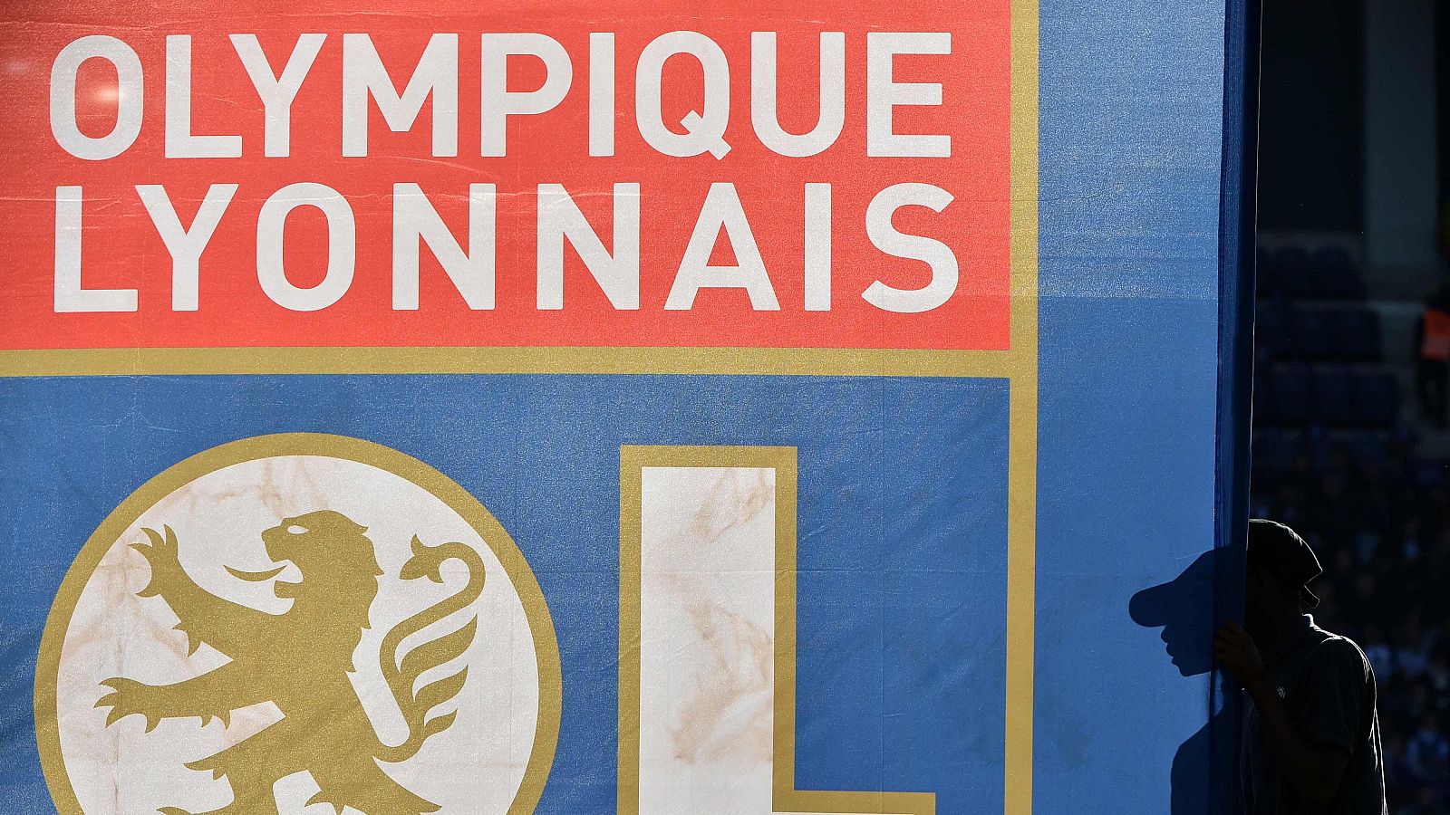 Gran escudo del Olympique Lyonnais en azul y rojo; nombre del equipo en blanco y león dorado en círculo blanco sobre azul. Silueta parcial de una persona a la derecha.