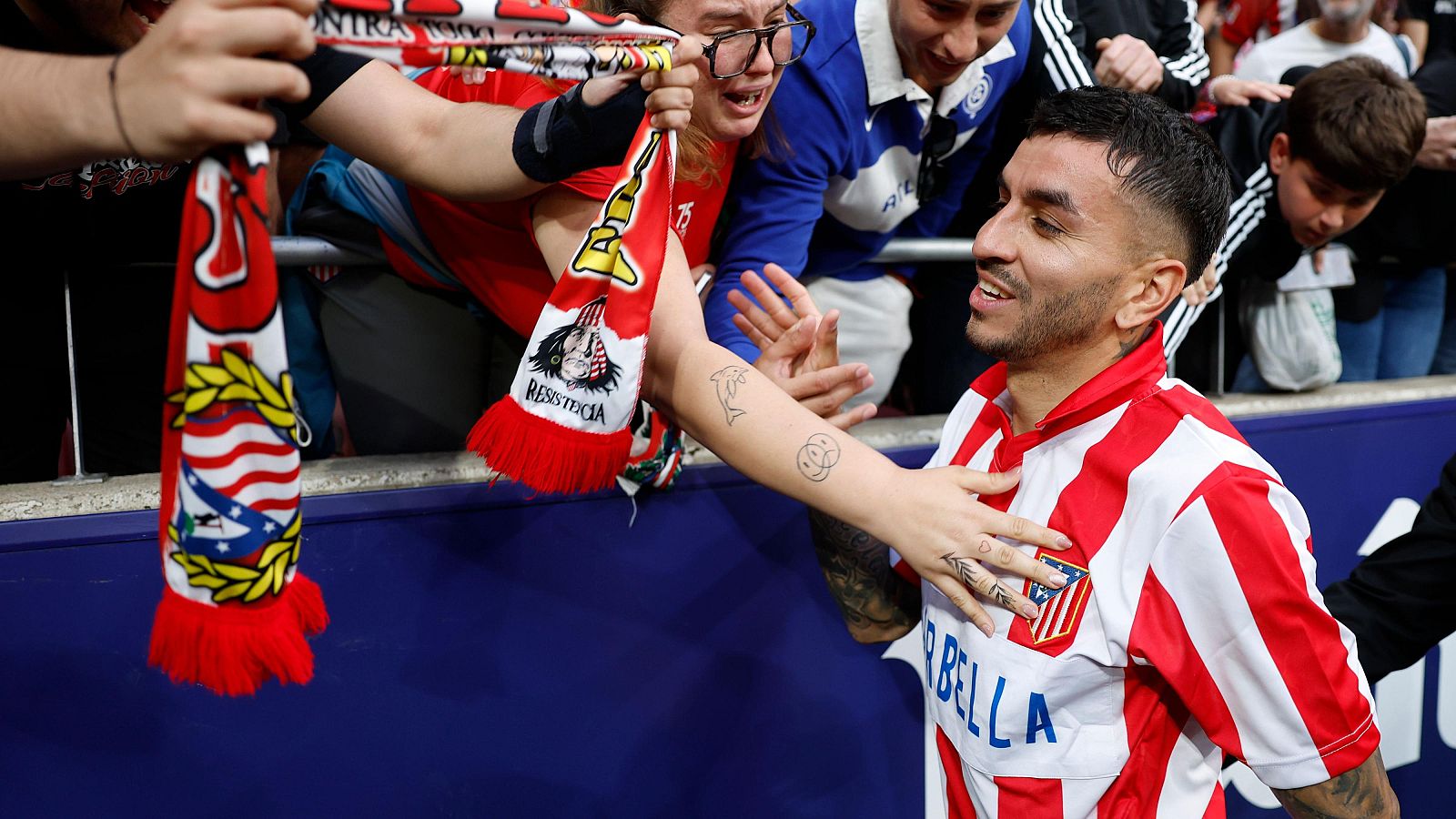 Despedida emotiva de Ángel Correa en el Metropolitano.  El jugador rojiblanco interactúa con aficionados, algunos con lágrimas, que le ofrecen bufandas. Se ven tatuajes en una mano que le toca.