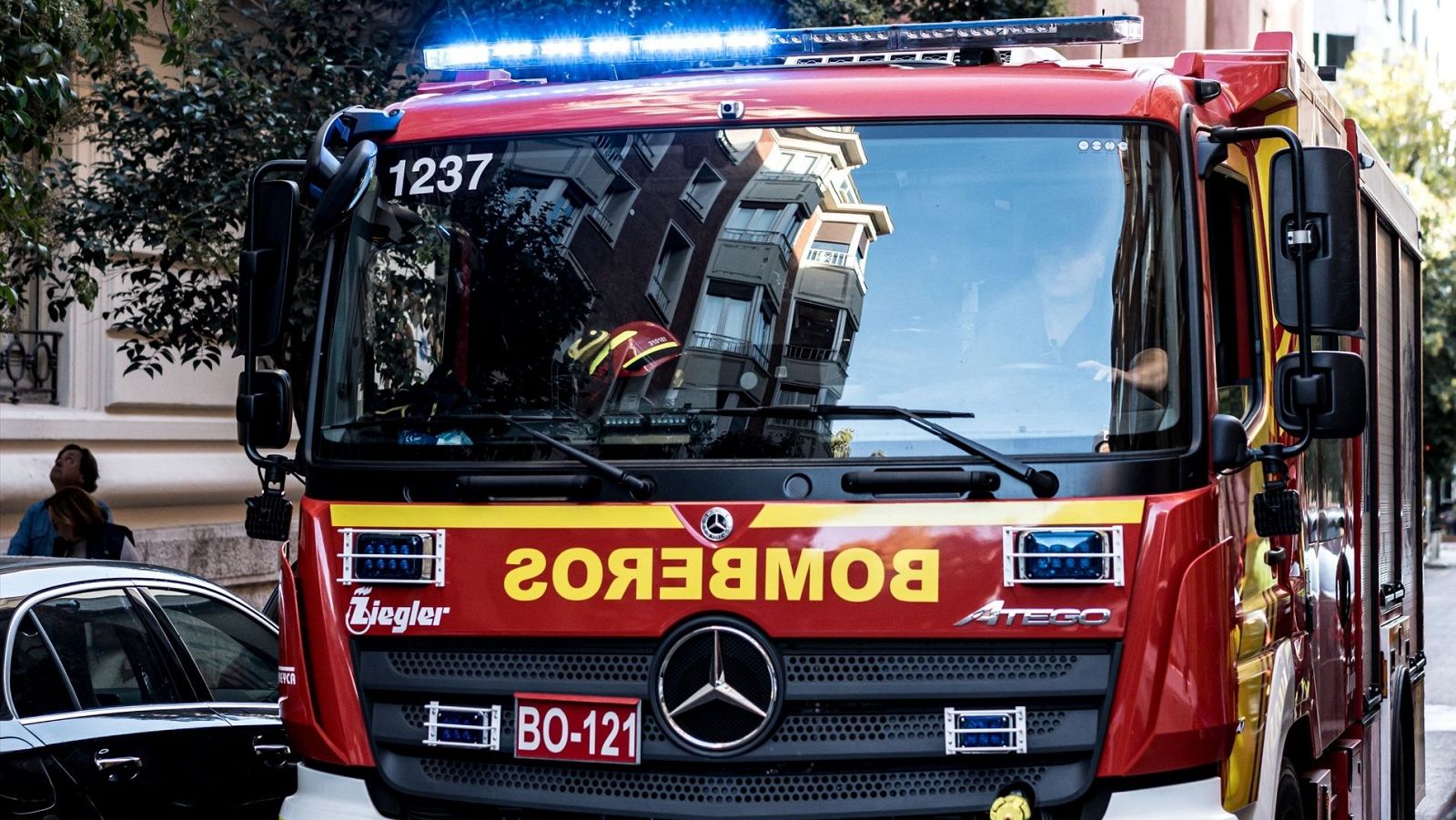 Camión de bomberos rojo Mercedes Atego (BO-121, 1237), luces azules activadas, 'BOMBEROS' reflejado en el parabrisas.  Coche negro y persona parcialmente visibles.