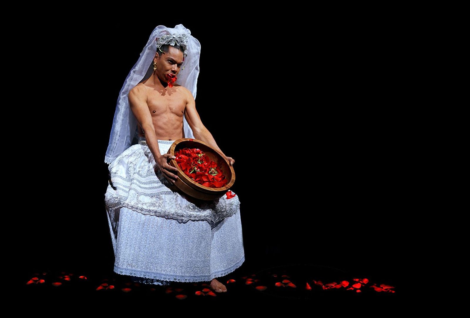Actor/bailarín en escena teatral con falda y velo blancos, sosteniendo un cuenco con pétalos rojos. Pétalos rojos cubren el suelo, creando un contraste con el vestuario blanco.