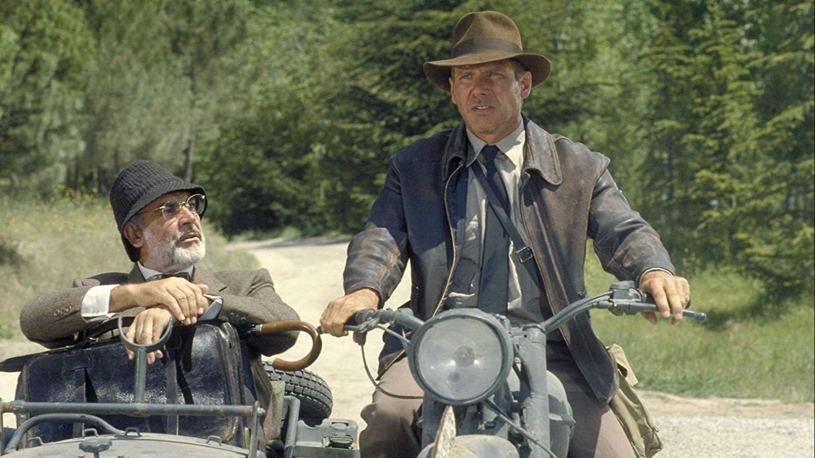 Indiana Jones (Harrison Ford) en motocicleta con sidecar, donde viaja acompañado por un hombre con traje y bastón (Sean Connery). Escena en camino de tierra arbolado.
