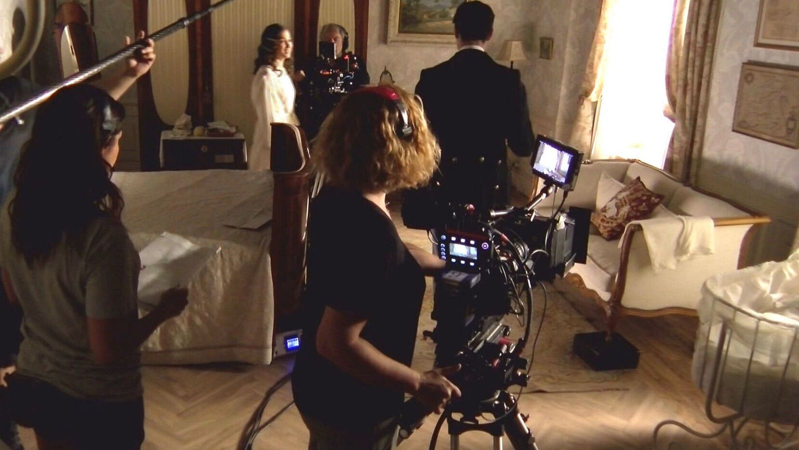 Rodaje de una escena de 'La Promesa': la actriz Carmen Asecas, vestida de época, es grabada por la operadora de cámara y su ayudante de dirección.  Se aprecia una iluminación cálida y un decorado de época.