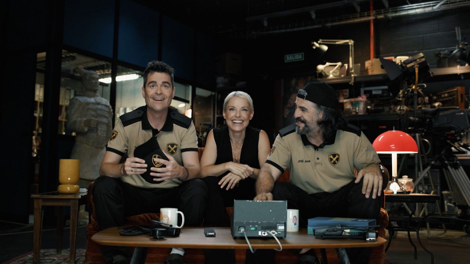 Tres personas sonrientes sentadas en un sofá de un plató de televisión, con objetos sobre una mesa baja y una estantería con material audiovisual al fondo.