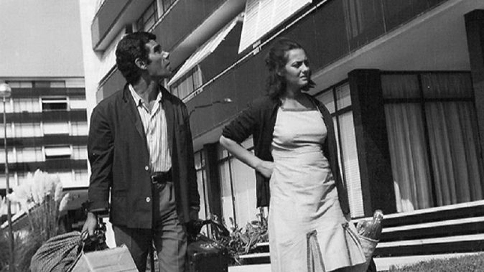 Pareja en calle frente a edificio moderno; escena en blanco y negro, posiblemente de mudanza o compra.