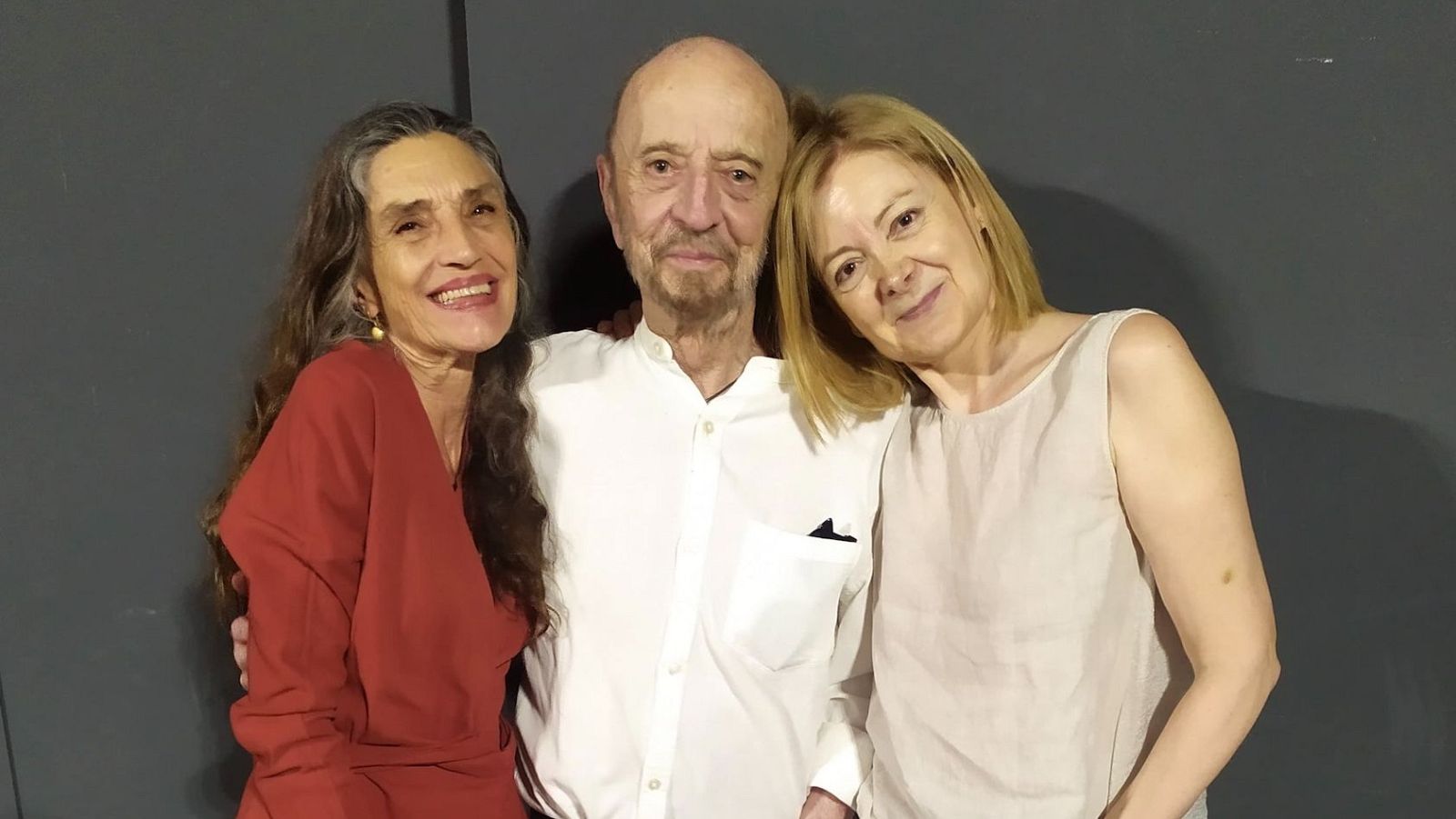 Tres personas posan juntas: una mujer con vestido rojo oscuro, un hombre con camisa blanca y otra mujer con blusa beige. Fondo gris oscuro.