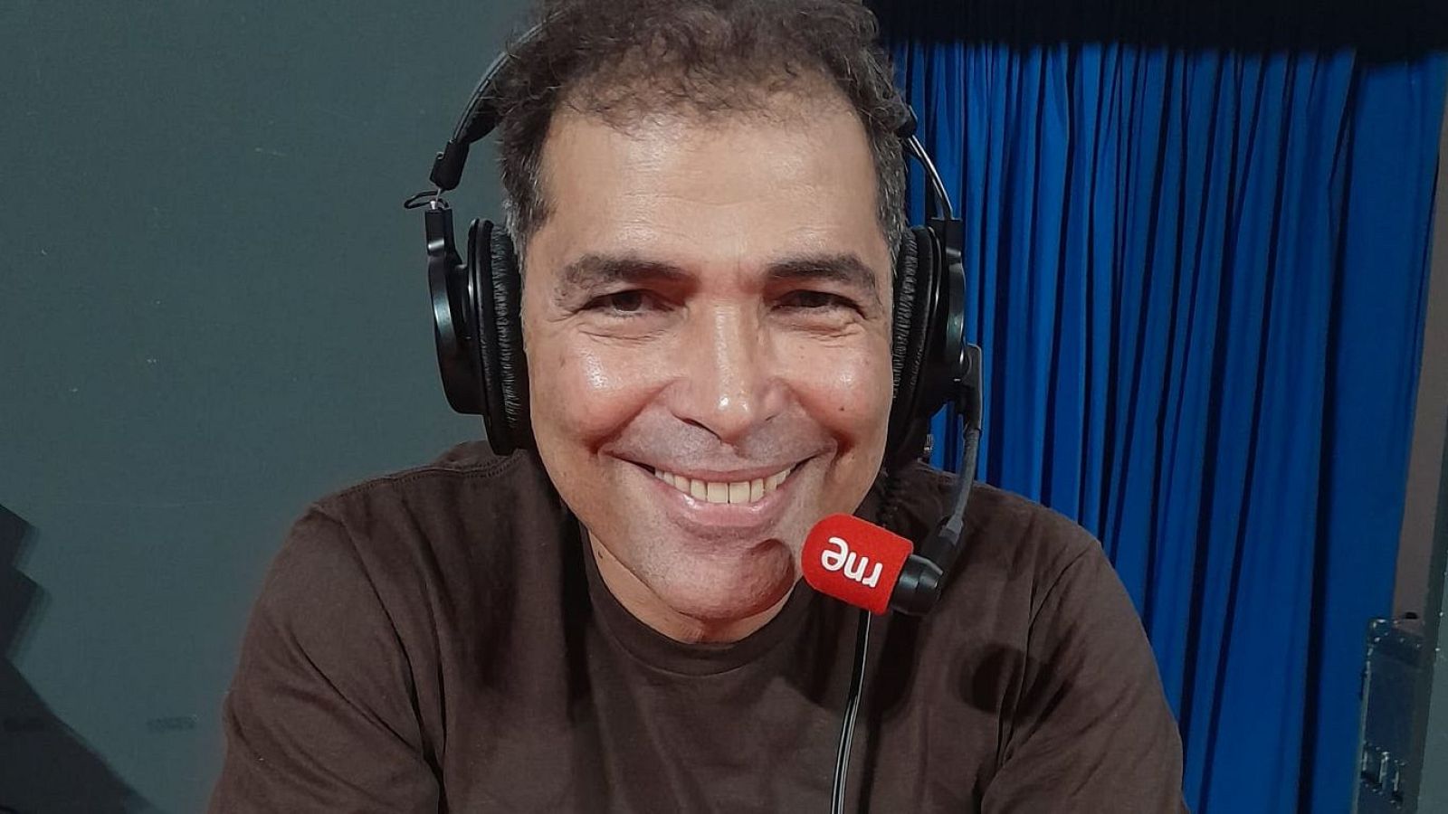 Actor y presentador cubano en estudio de radio con auriculares y micrófono de RNE, sonriendo a cámara.