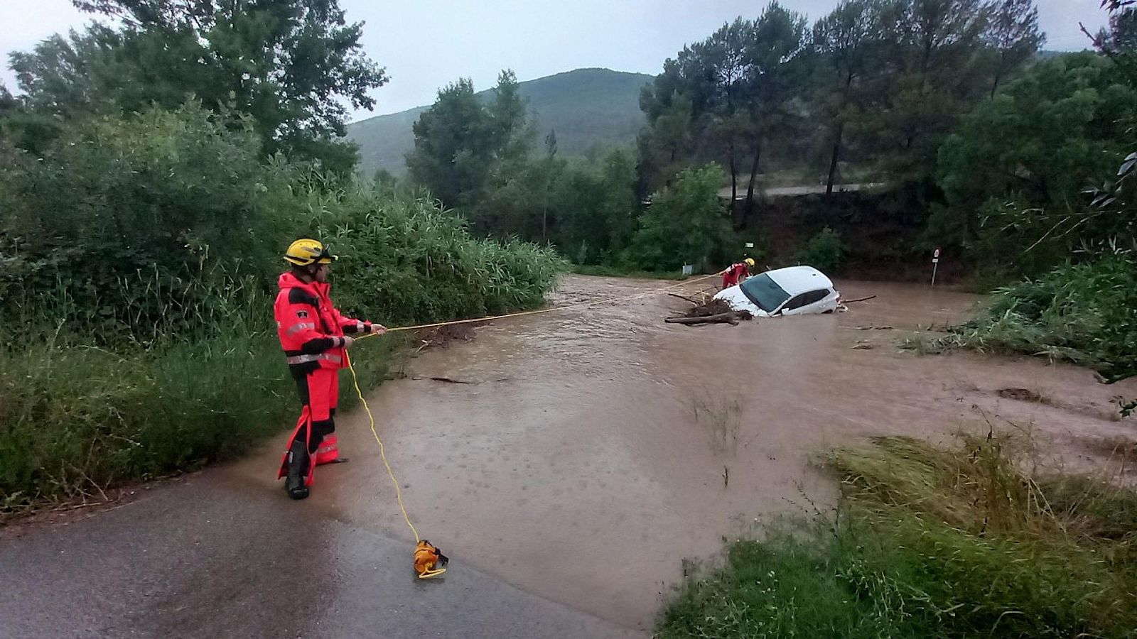 Rescate de un coche blanco parcialmente sumergido en un río por dos bomberos que usan una cuerda amarilla. Entorno rural tras una aparente inundación.