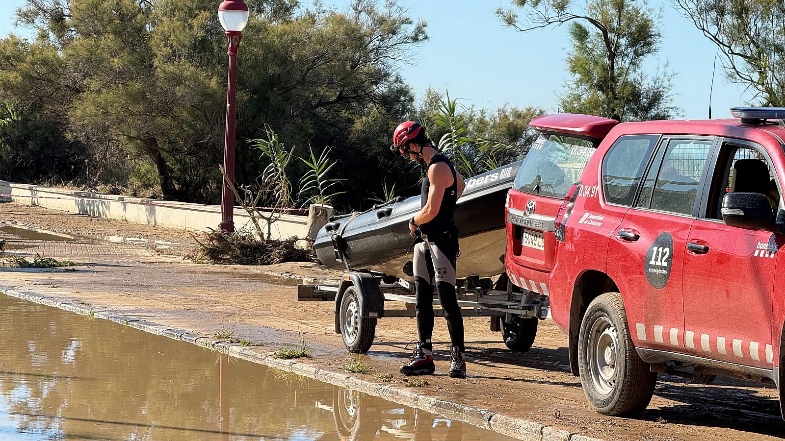 Bombero equipado para intervención subacuática junto a lancha neumática en remolque enganchado a un todoterreno rojo con el número 112 y la palabra "Bombers". Entorno inundado.