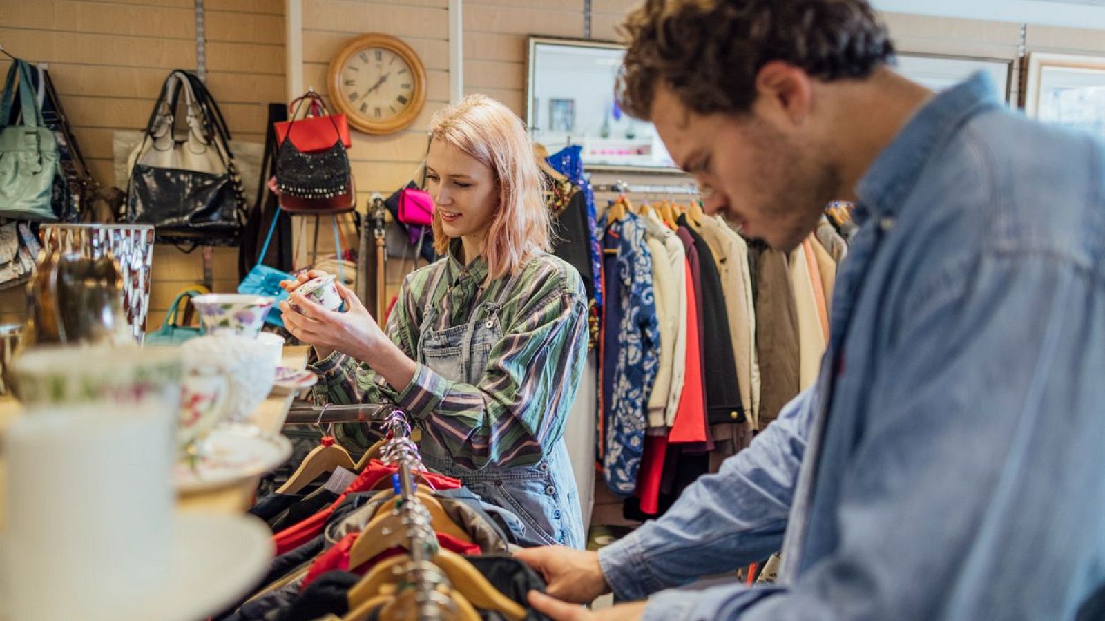 Pareja o amigos revisan artículos en una tienda de segunda mano; ella observa vajilla, él ropa.  Ambiente vintage con bolsos y estanterías.