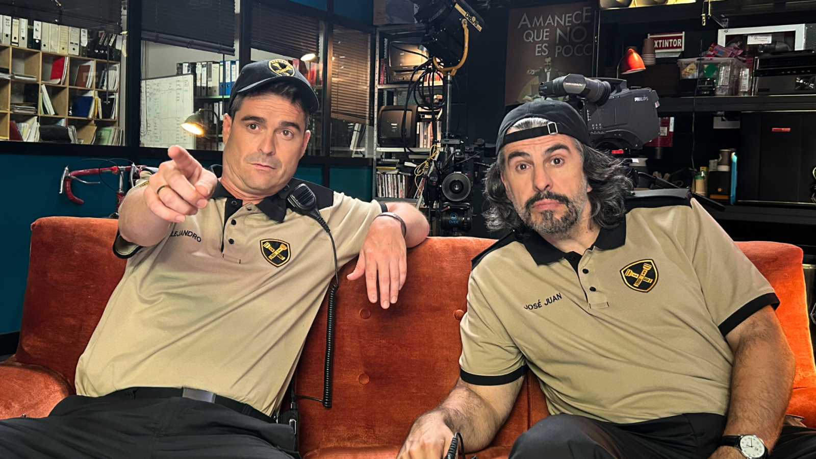 Dos hombres en un sofá de plató de TV, con polos y gorras, uno señala a cámara.  Se ve equipo de grabación y un cartel con una frase.