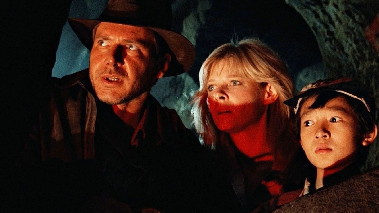 Tres personajes, con expresiones de alerta y miedo, observan algo fuera de cuadro en una cueva oscura con iluminación rojiza.