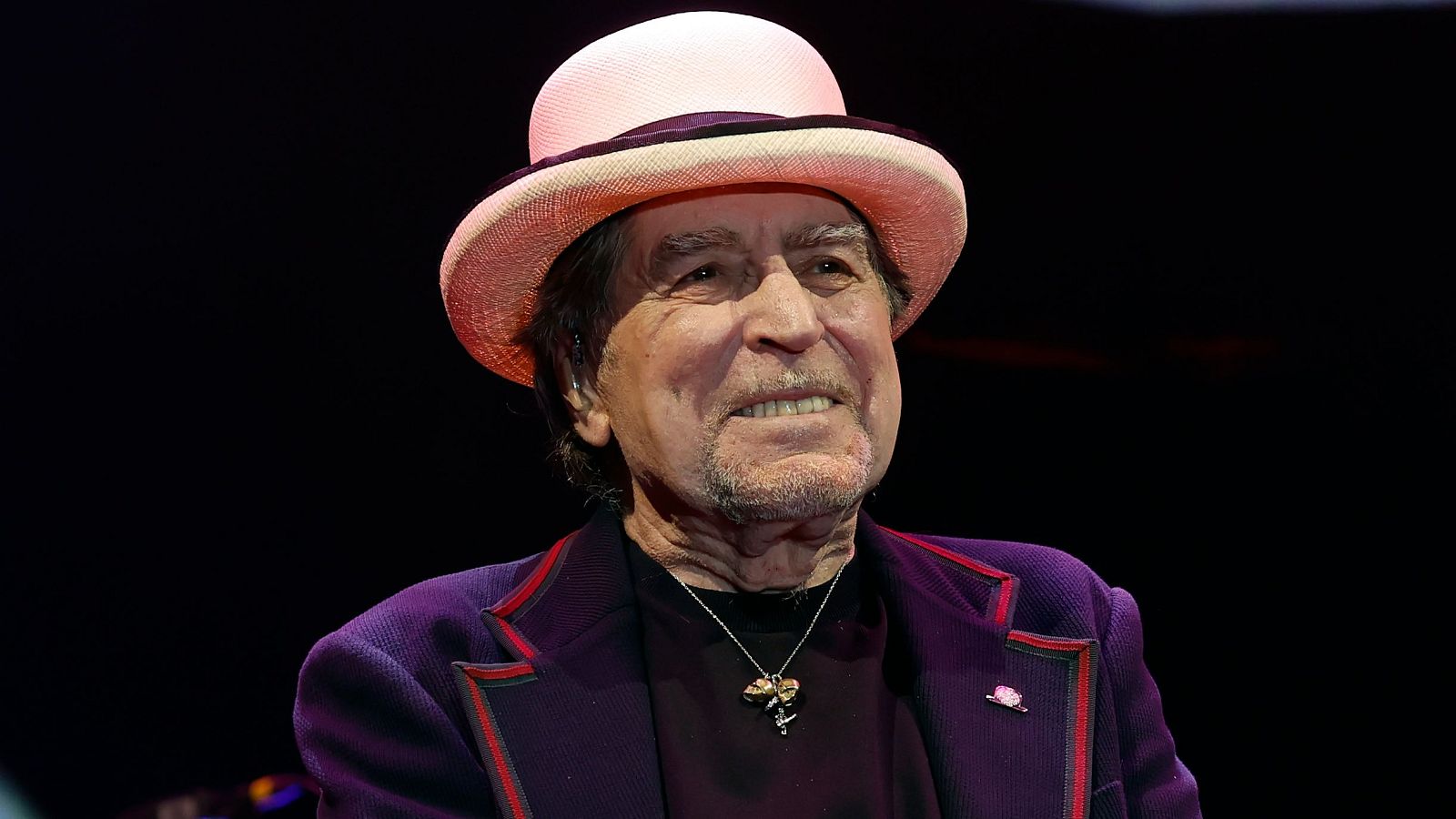 Joaquín Sabina en concierto: sombrero rosa, chaqueta morada con ribetes rojos, camiseta oscura y expresión serena. Fondo oscuro.