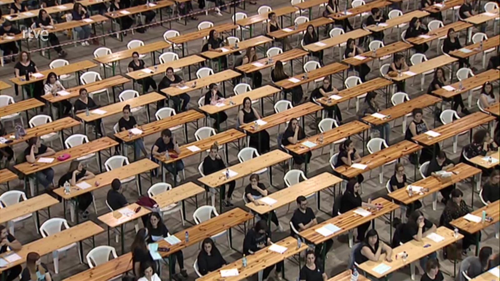 Vista aérea de una sala llena de opositores realizando un examen; mesas individuales con materiales, ambiente de concentración. Logo RTVE visible.