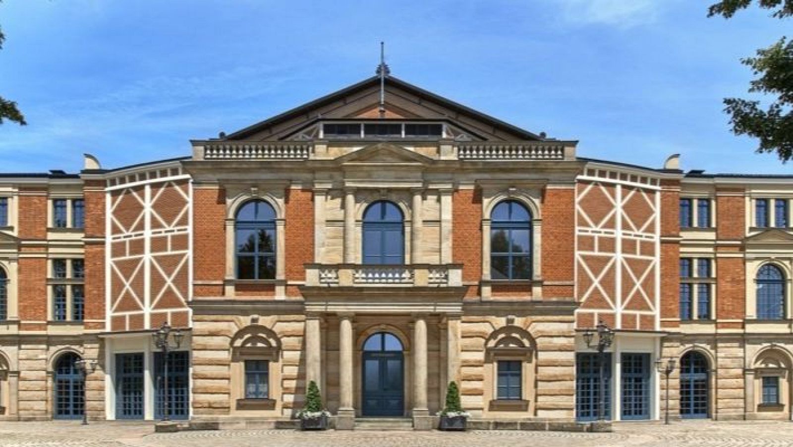 Fachada del Bayreuther Festspielhaus: ladrillo rojo y piedra clara, detalles decorativos en 'X', entrada principal con columnas y puerta azul oscura, ventanas arqueadas y tejado oscuro.