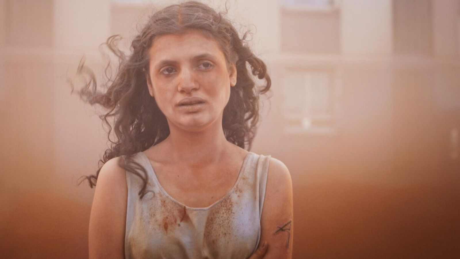 Joven con cabello oscuro y rostro sucio, mirada seria, en un entorno polvoriento y borroso.  Imagen de alta intensidad, posiblemente de una escena de conflicto o supervivencia.