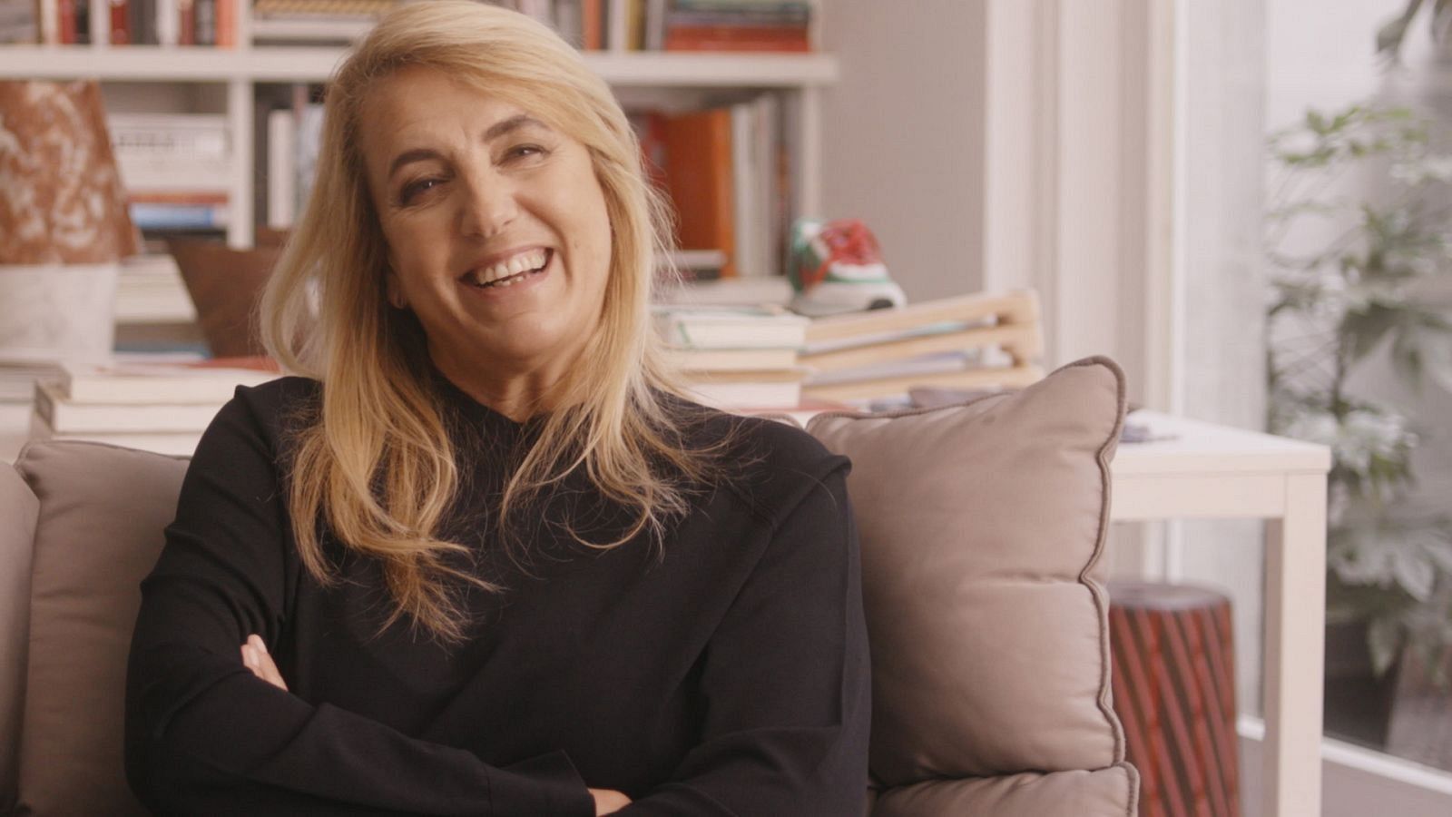 Diseñadora sentada en un sofá claro, sonriendo a cámara con los brazos cruzados y blusa negra.  Ambiente tranquilo con libros y objetos decorativos de fondo.
