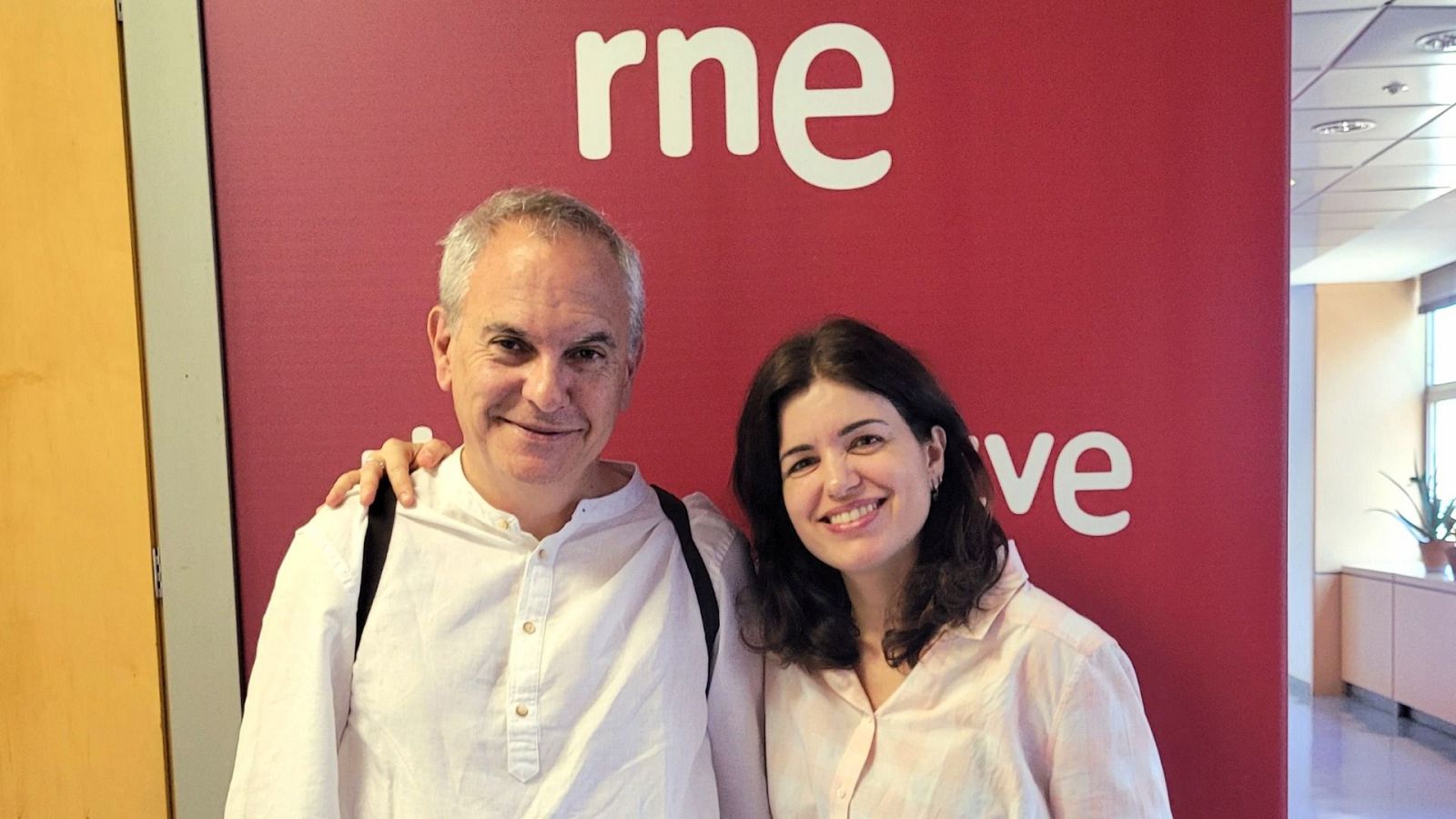 Fotografía de dos personas sonriendo junto a un cartel de Radio Nacional de España.  Pareja en un pasillo, posiblemente promocional de un curso de meditación.