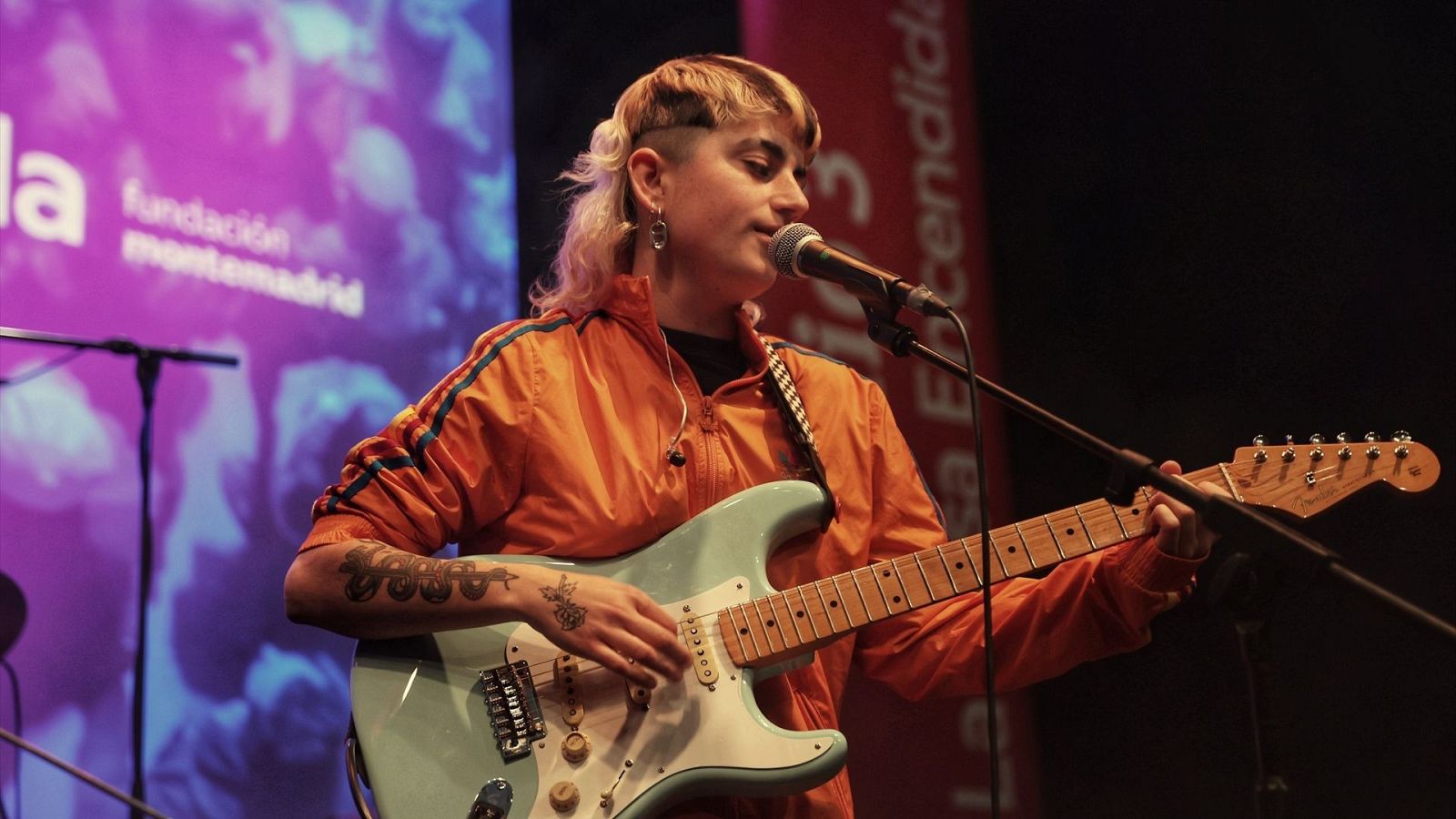 Cantante con corte mullet rubio y chaqueta naranja tocando guitarra verde claro en concierto.  Se observa un logo de la Fundación Montemadrid y parte de un cartel rojo en el fondo.