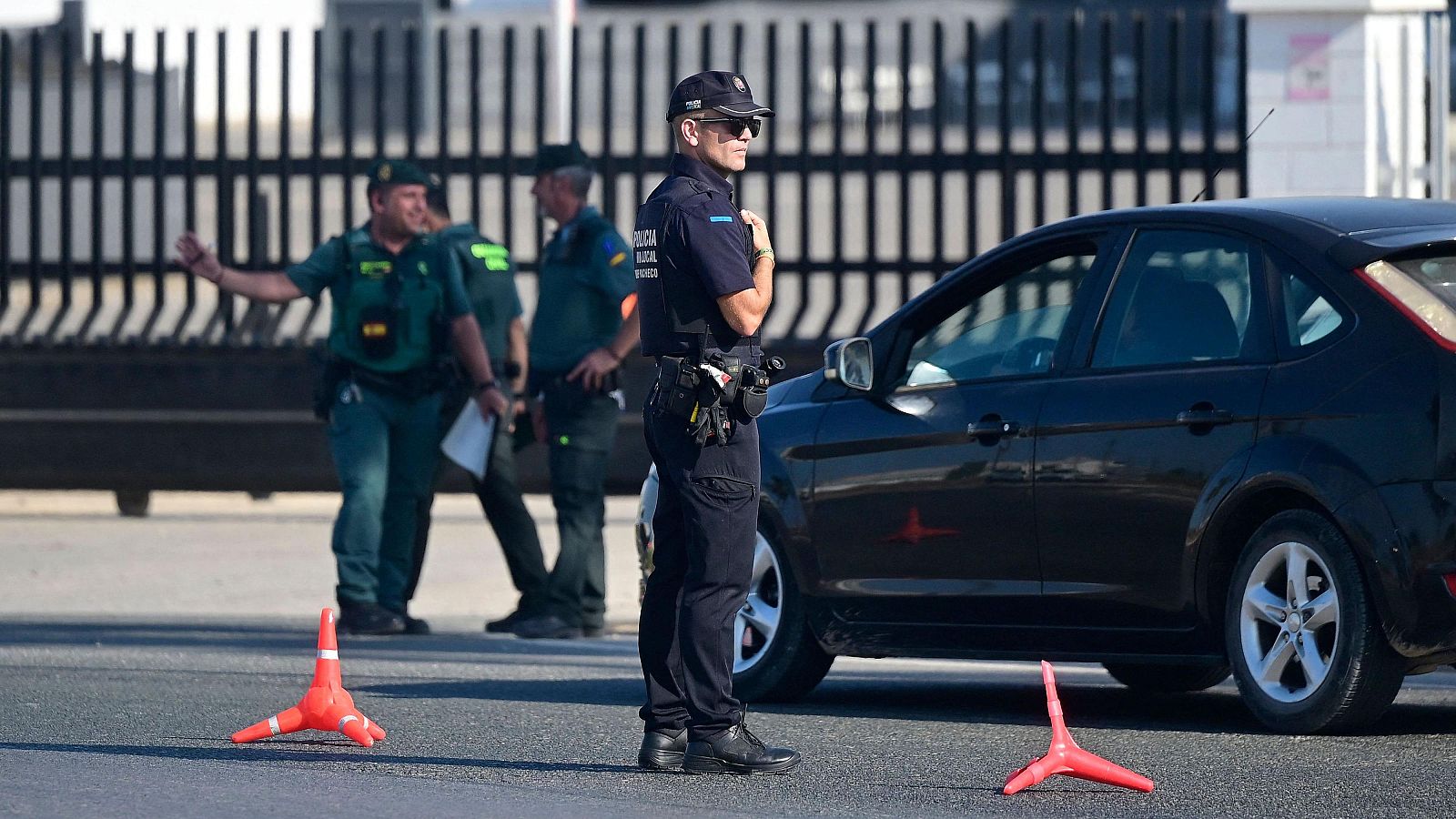 Agente de policía en uniforme oscuro y gafas de sol, vigilando una zona con presencia de otros agentes y conos de tráfico.