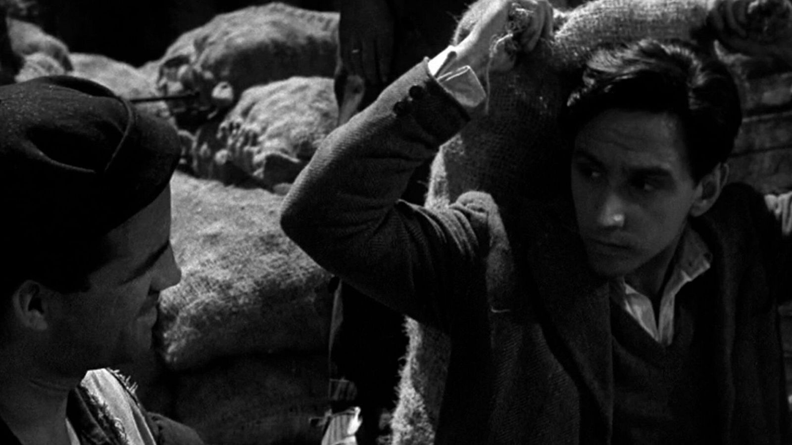 Imagen en blanco y negro de dos hombres en un almacén, uno con un saco grande.  Ambientación de trabajo físico y precariedad.