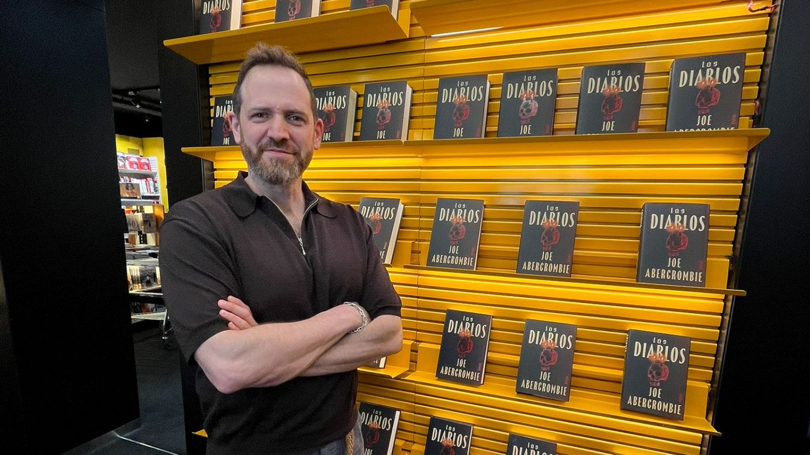 Joe Abercrombie de pie, con los brazos cruzados y una sonrisa, delante de una estantería amarilla llena de copias del libro Los Diablos
