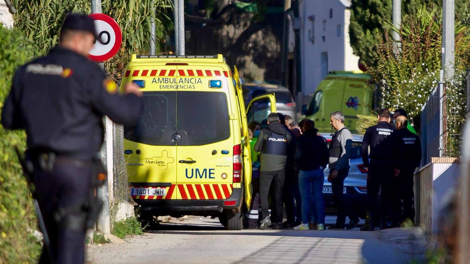 Accidente con ambulancia en calle residencial.  Personal sanitario atiende a pacientes.  Policía y civiles presentes. Se observa señal de límite de velocidad.