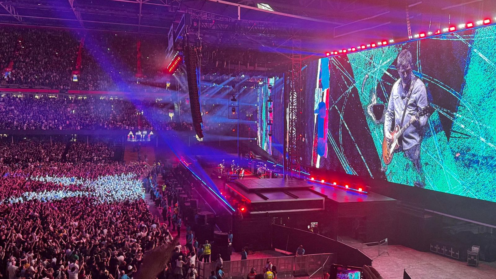 Estadio abarrotado en un concierto de Oasis.  Gran pantalla muestra a un guitarrista tocando.  Ambiente vibrante y festivo.