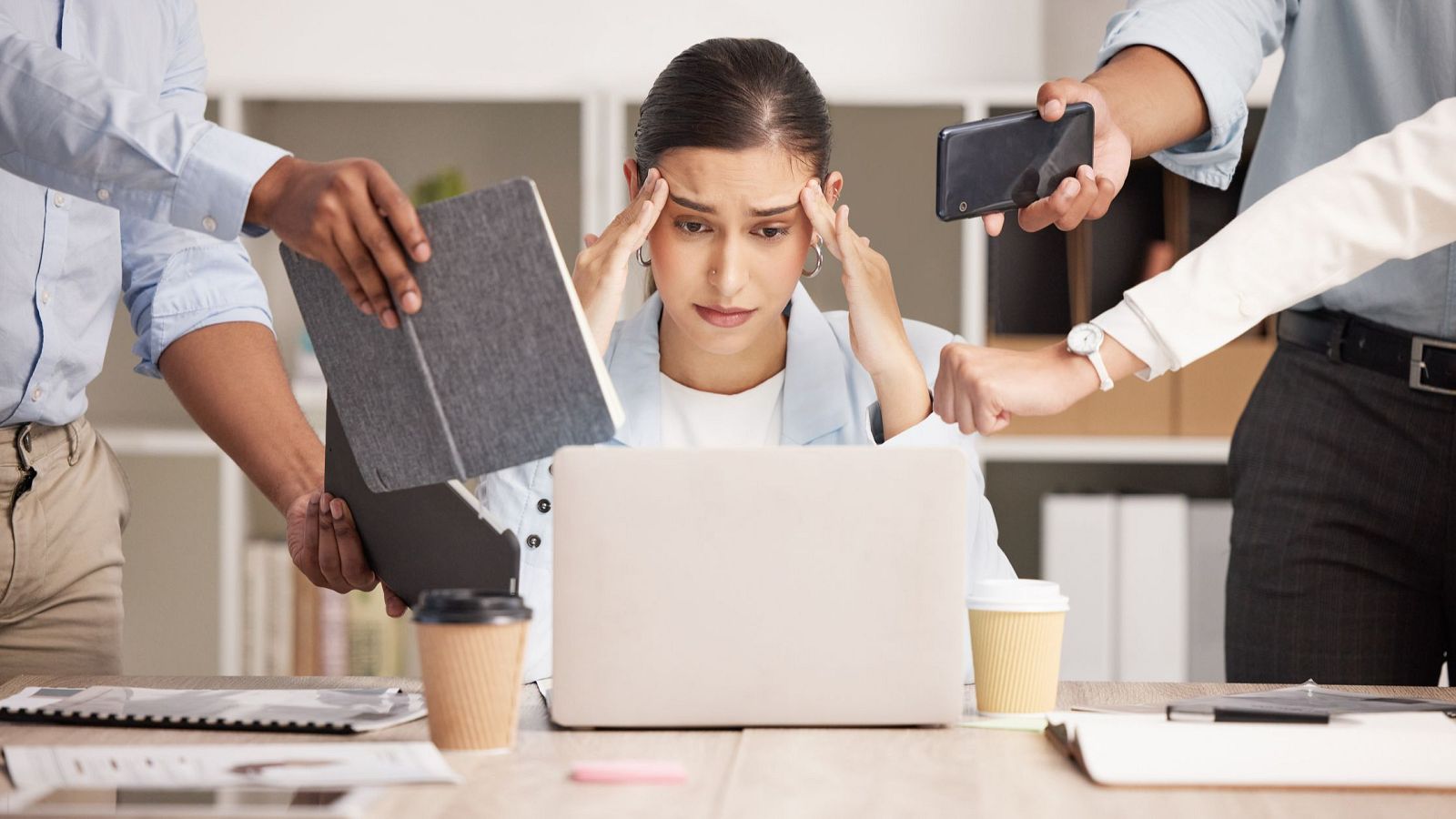 Mujer estresada en su escritorio, rodeada de manos que simbolizan la presión laboral y la sobrecarga de tareas.  Imagen que representa el síndrome del trabajador quemado.