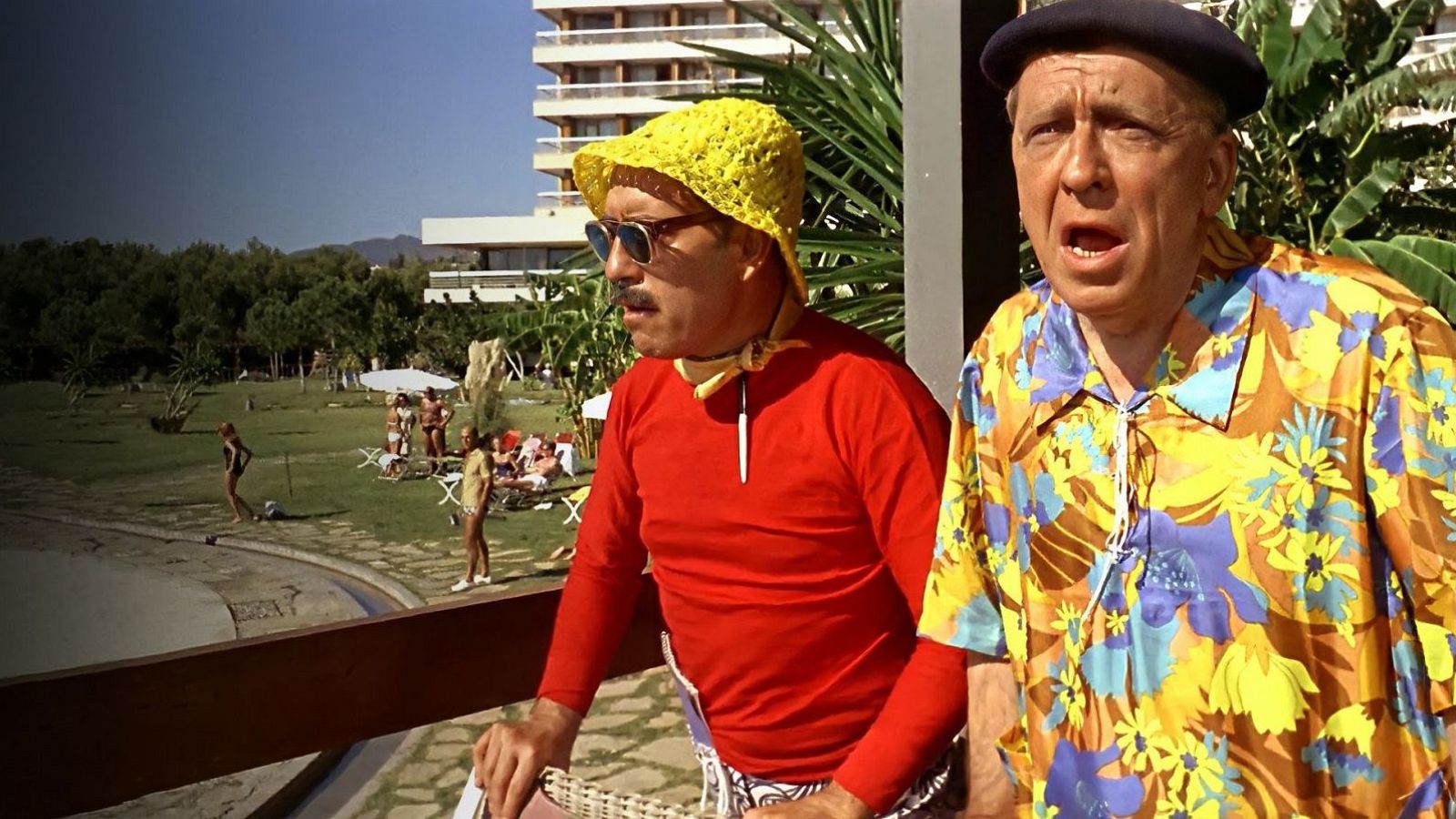 Dos actores, uno con sombrero amarillo y otro con boina azul, miran sorprendidos en una terraza con piscina al fondo. Promoción de cine español.