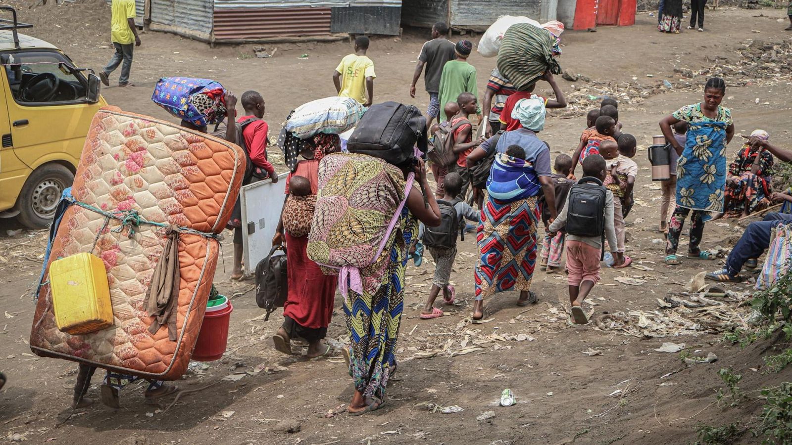 Refugiados, mayormente mujeres y niños, transportan sus pertenencias por un camino de tierra, mostrando un entorno de campamento improvisado.