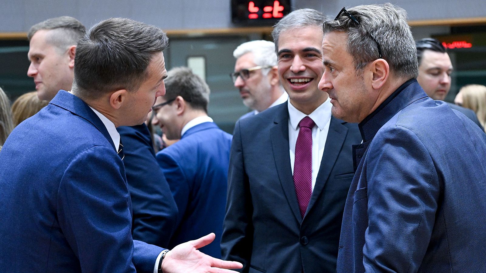 Reunión de ministros europeos (Polonia, UE y Luxemburgo) en Bruselas. Conversación entre tres hombres en traje durante un evento oficial, con un marcador electrónico visible.