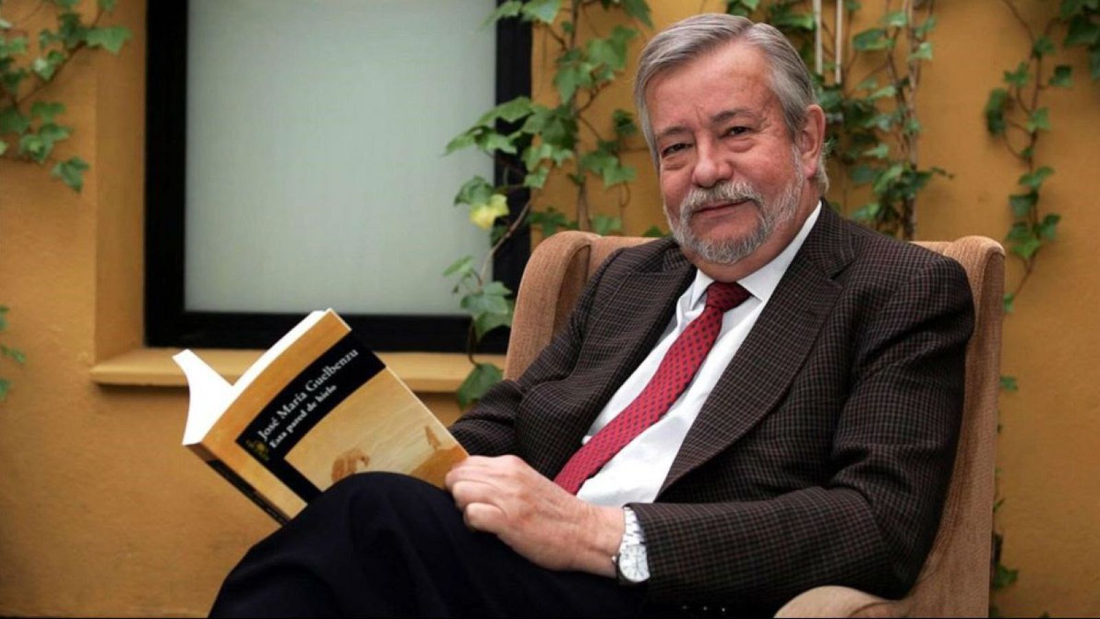 Escritor mayor con traje oscuro y corbata roja, sentado, sosteniendo su libro "Esta paz de la lluvia". Fondo con pared ocre y plantas.